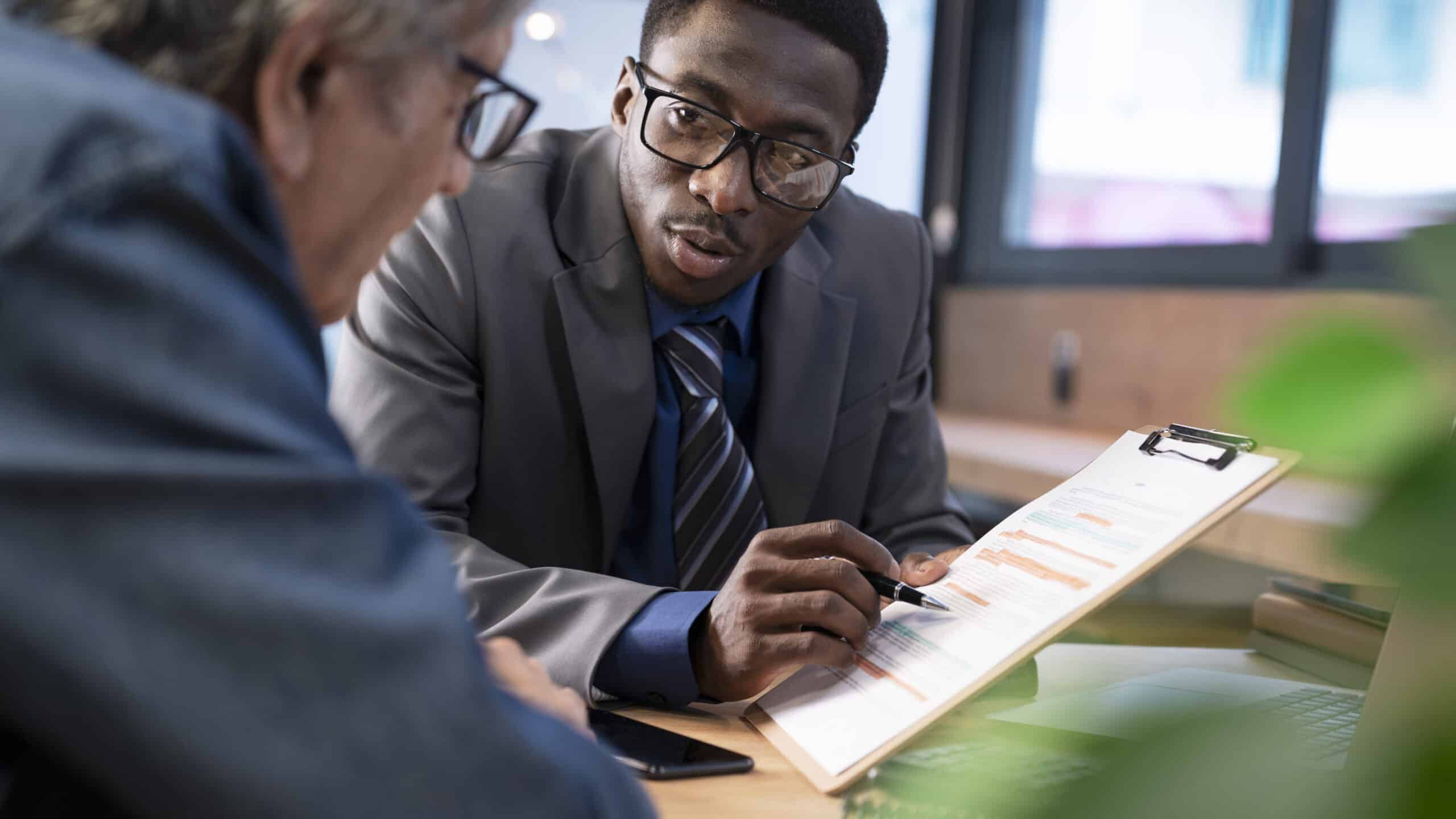 Two professionals review a clipboard with a resume or report, one guiding with a pen in a business discussion at a desk