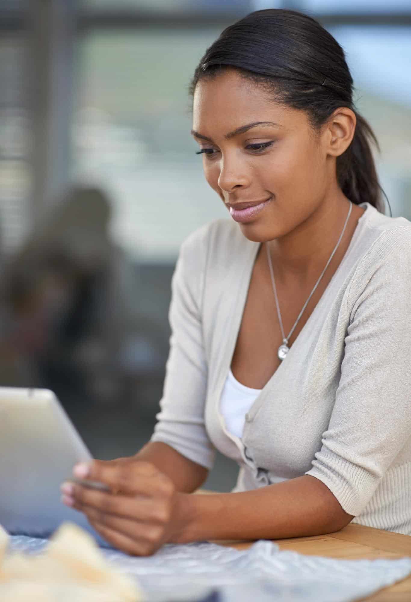 Woman in a light cardigan sits at a desk, typing on a laptop while holding a smartphone.