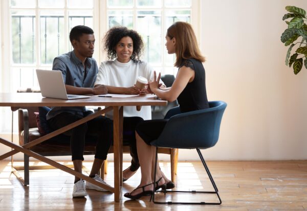 Three people sit around a wooden table in a bright room, having a discussion; one holds a coffee cup while another gestures with her hands.